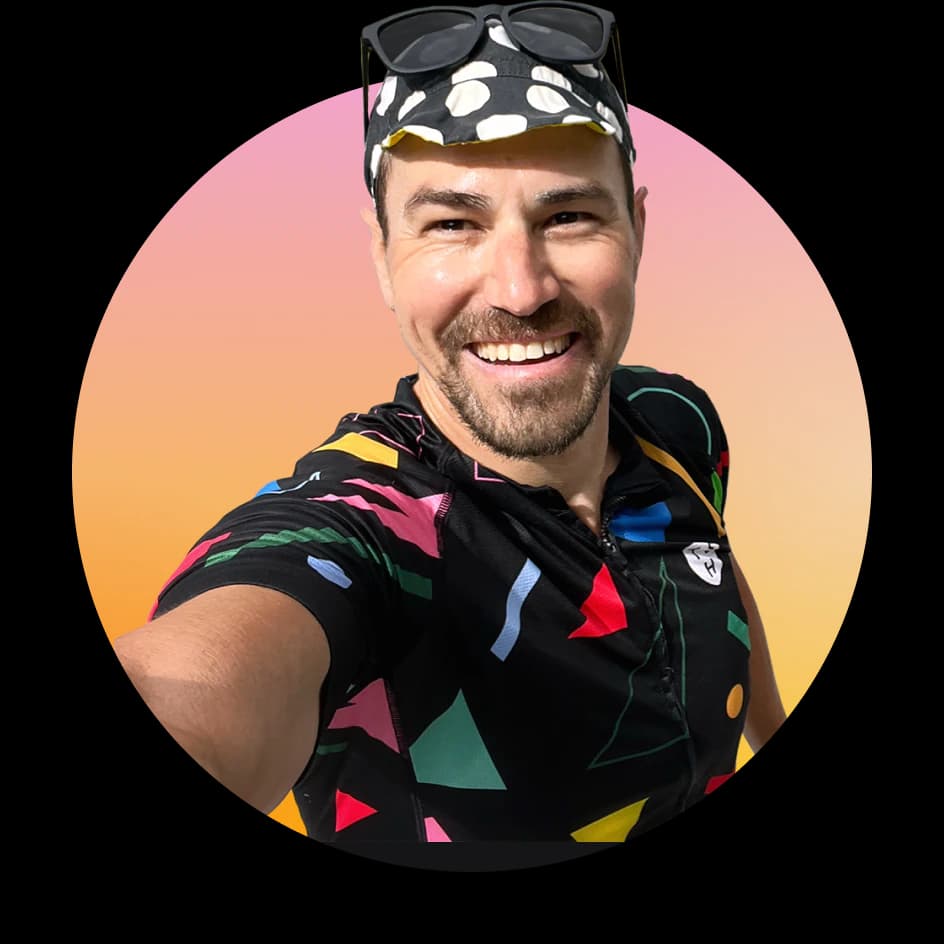 Blair Metcalf grinning, in a polka-dot cycling cap with sunglasses pushed up on it and a black jersey scattered with bright shapes. The photo is a disc with a sunset gradient behind him, and the cap breaks out of the top of it.