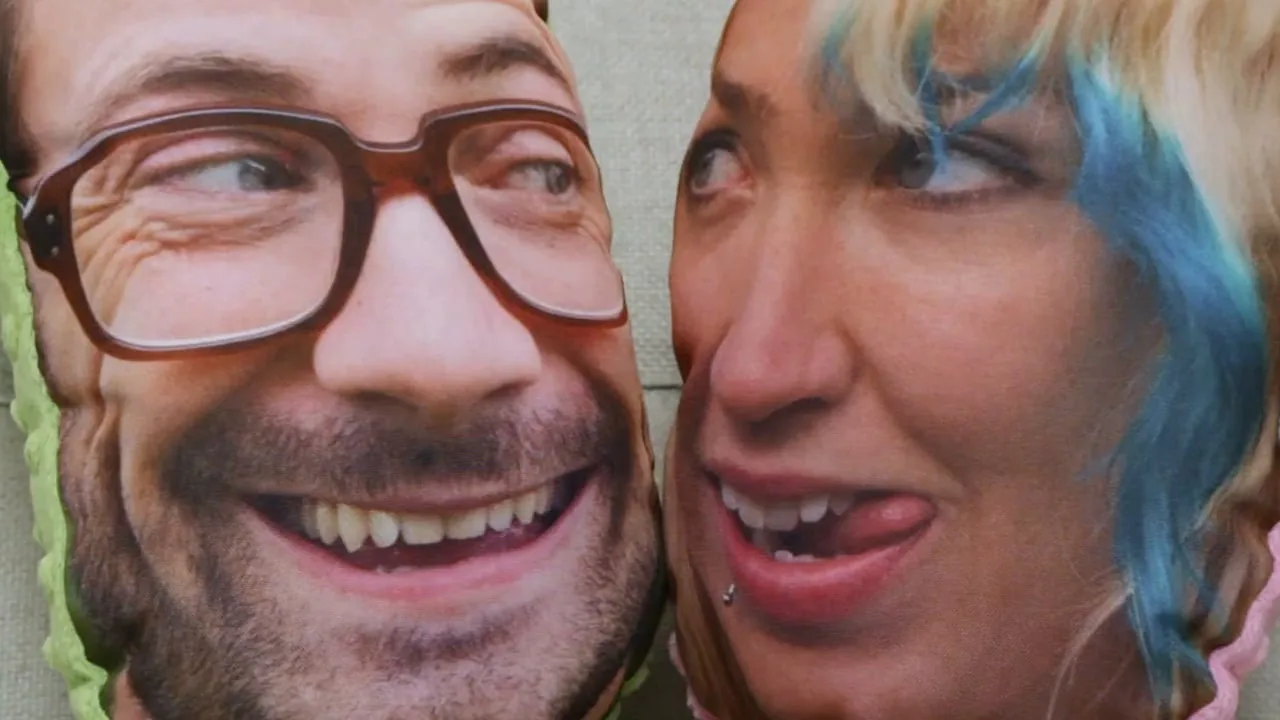 Two face pillows pressed cheek to cheek in close-up: a grinning man in thick-rimmed glasses, and a woman with blue hair sticking out her tongue.